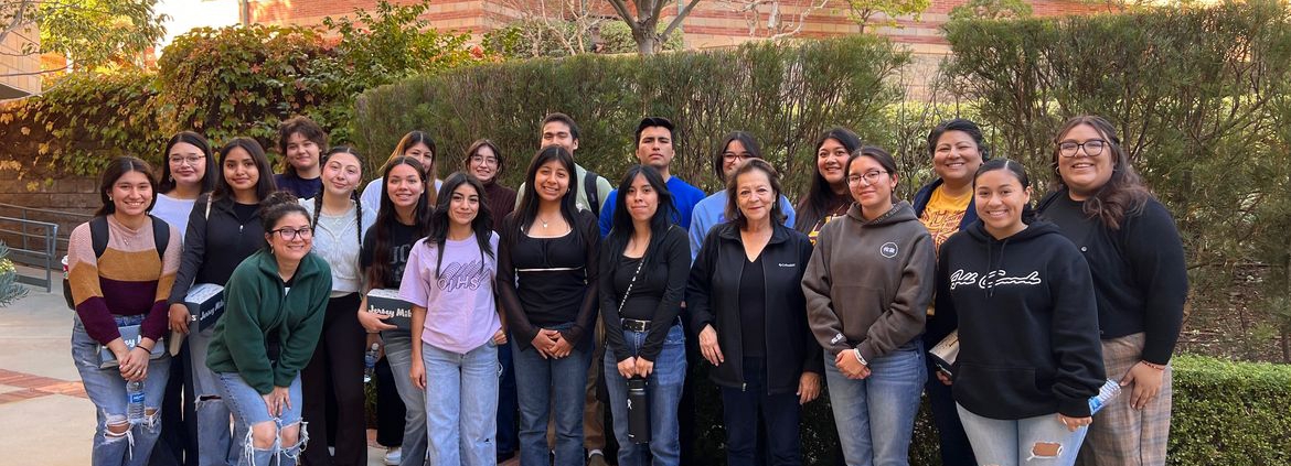 A group of students pose for a photo with bushes in the background on UCLA's campus.