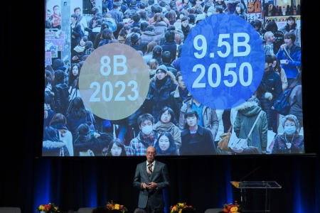 Jack Bobo, a man wearing a grey suit stands on stage in front of a screen that shows a crowd of people with the estimated population totals: 8 billion 2023 and 9.5 billion in 2050.