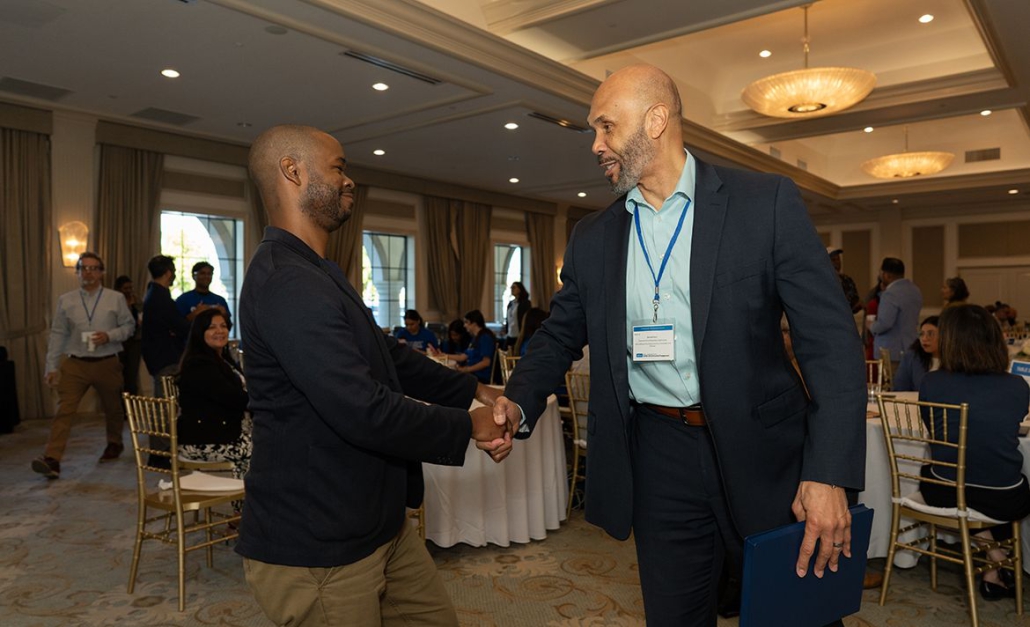 Two men standing in a ballroom shake hands.