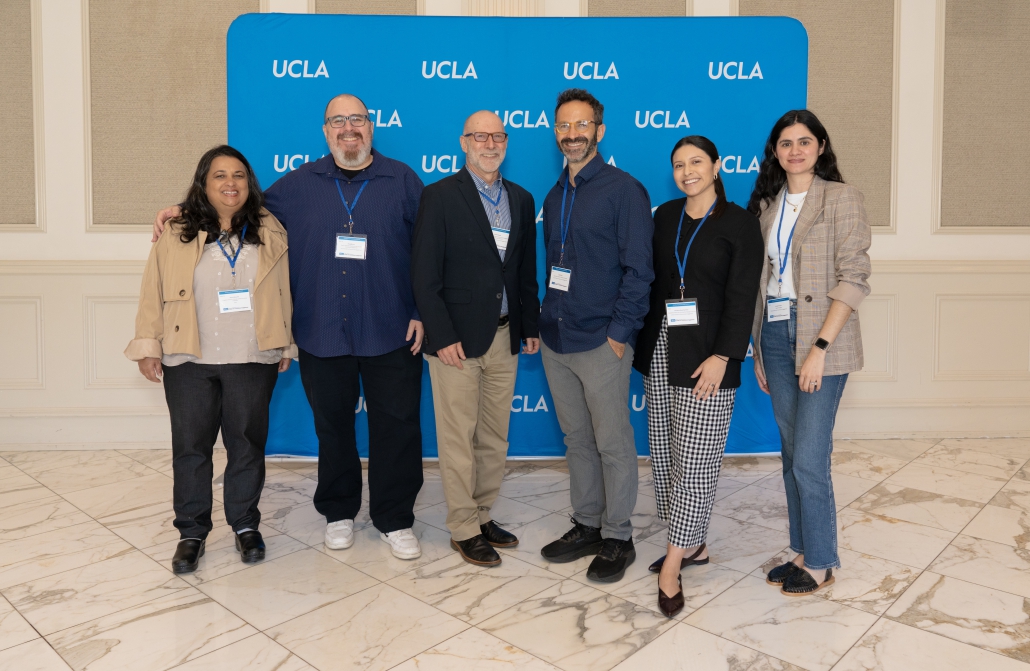A group of six people stand in front of a step and repeat with the UCLA logo on it.