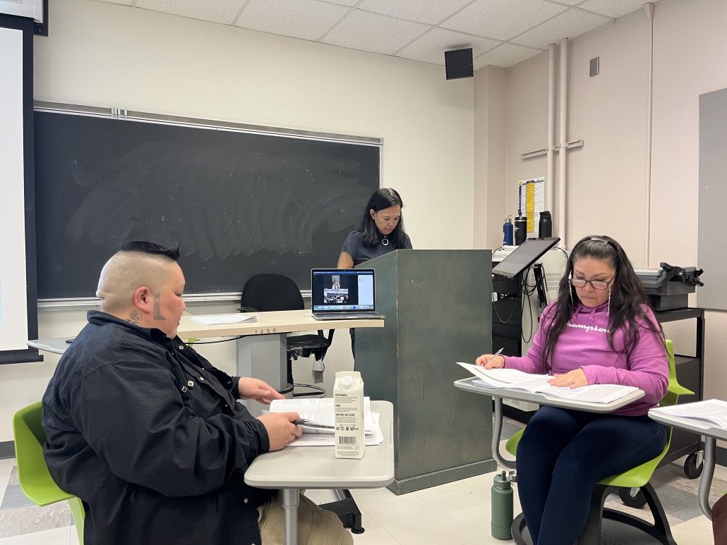 Three women in a classroom. One standing at a podium and the other two are sitting down reading.