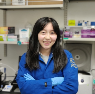 A photo of a young woman with brown hair standing in a lab wearing a blue lab coat and gloves.