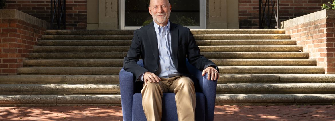 Shalom Staub in light-blue button-down, dark jacket and tan pants, seated in a blue armchair in front of a brick building.