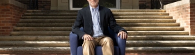 Shalom Staub in light-blue button-down, dark jacket and tan pants, seated in a blue armchair in front of a brick building.
