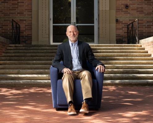 Shalom Staub in light-blue button-down, dark jacket and tan pants, seated in a blue armchair in front of a brick building.