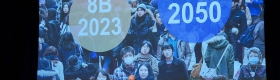 A man in a suit stands in front of a large screen showing a power point presentation that features a large group of people and text that conveys that there are 8 billion people in 2023 and 9.5 billion by 2050.