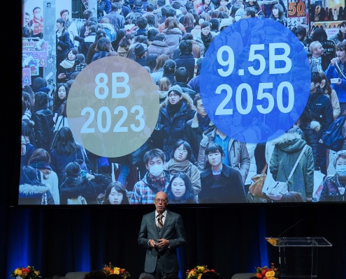 A man in a suit stands in front of a large screen showing a power point presentation that features a large group of people and text that conveys that there are 8 billion people in 2023 and 9.5 billion by 2050.