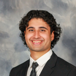 A man with dark curly hair wearing a suit and tie poses in front of a grey portrait studio background.