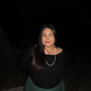 A woman with long dark hair wearing a white necklace, black shirt, and green pants stands in front of a black background.