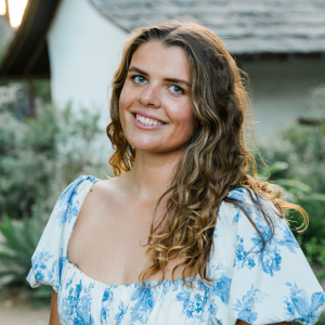 A woman with light brown hair wearing a white dress with blue florals stands outside and the background is out of focus.
