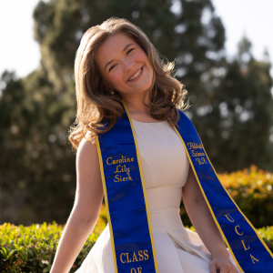 A woman with light brown hair stands outside with a tree in the distance. She wears a white dress and her UCLA graduation sash that says 