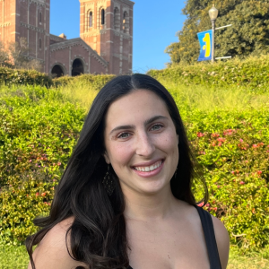A woman with long dark hair stands in front of a green bush. In the distance you can see UCLA's Royce Hall.