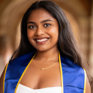 A woman with long dark hair wearing a white dress and her UCLA graduation sash smiles and looks directly at the camera.