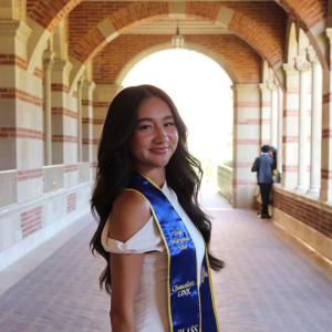A woman with long dark hair wearing a white dress and graduation sash stands under a brick arch. 