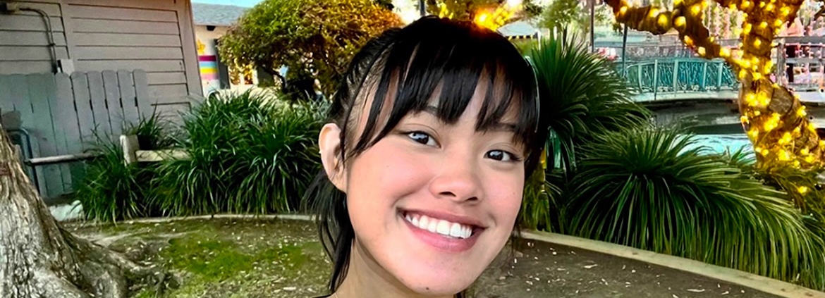 UCLA student Deanna Placa smiles for a selfie outdoors, surrounded by trees and greenery decorated with string lights.