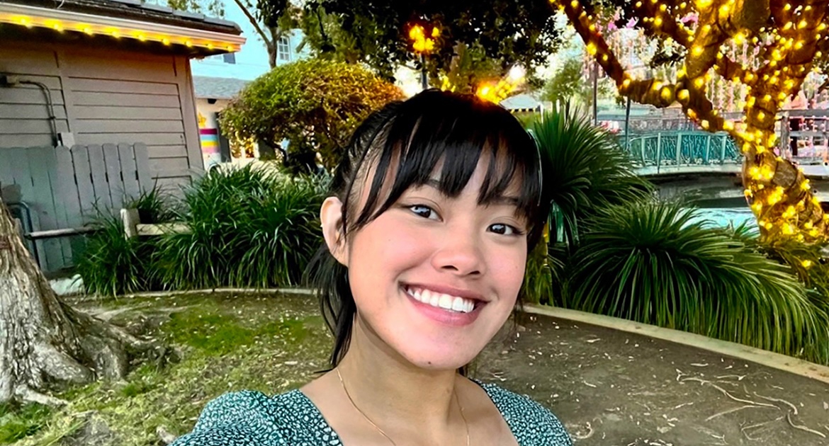 Deanna Placa-Newman Fellowship UCLA student Deanna Placa smiles for a selfie outdoors, surrounded by trees and greenery decorated with string lights.