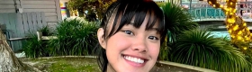 UCLA student Deanna Placa smiles for a selfie outdoors, surrounded by trees and greenery decorated with string lights.