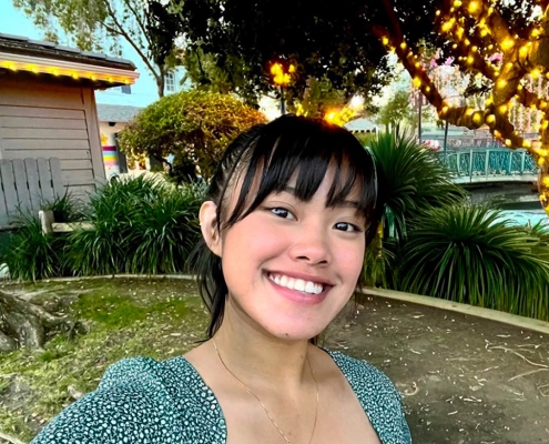 UCLA student Deanna Placa smiles for a selfie outdoors, surrounded by trees and greenery decorated with string lights.