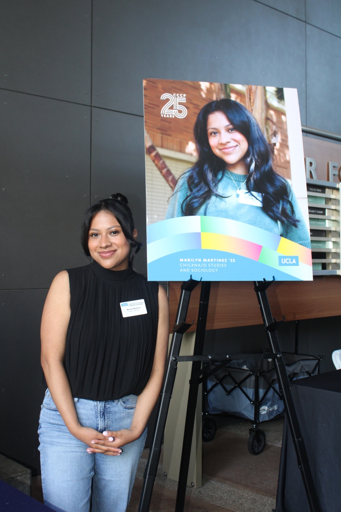 Marilyn Martinez, wearing a black top, jeans, and a name tag, stands next to a banner displaying her portrait and degree information.
