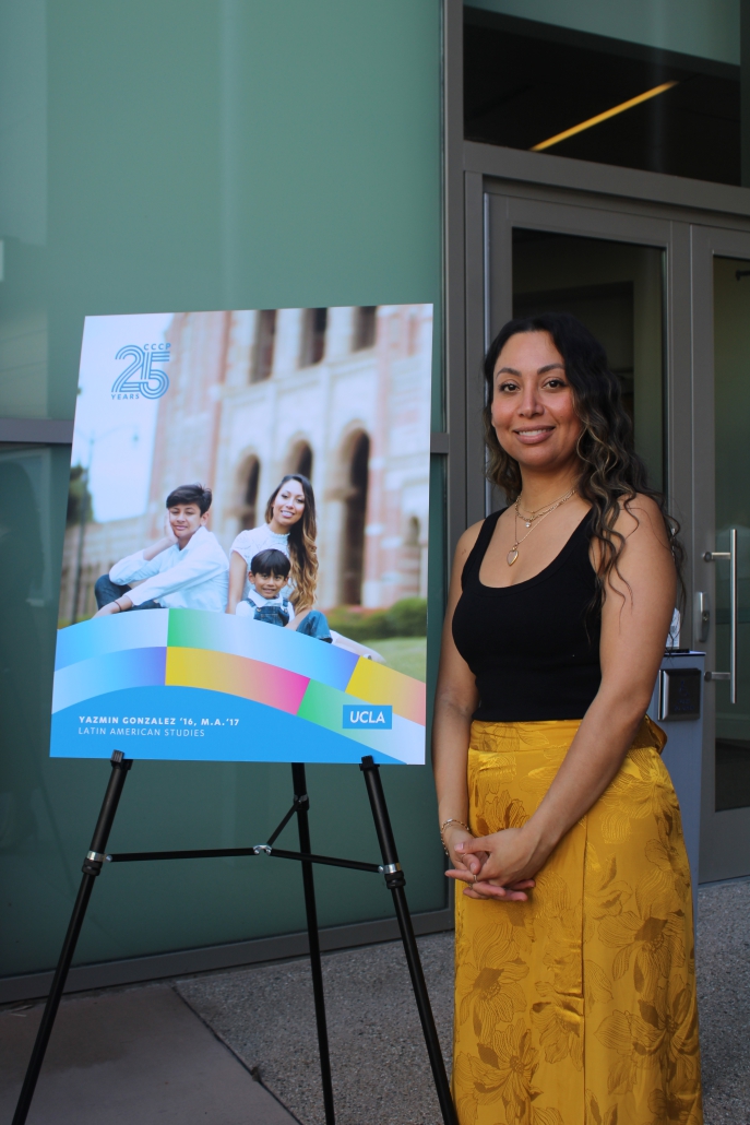 Yazmin Gonzalez, dressed in a black top and yellow patterned skirt, stands next to a poster on an easel highlighting her as a CCCP and UCLA alum.