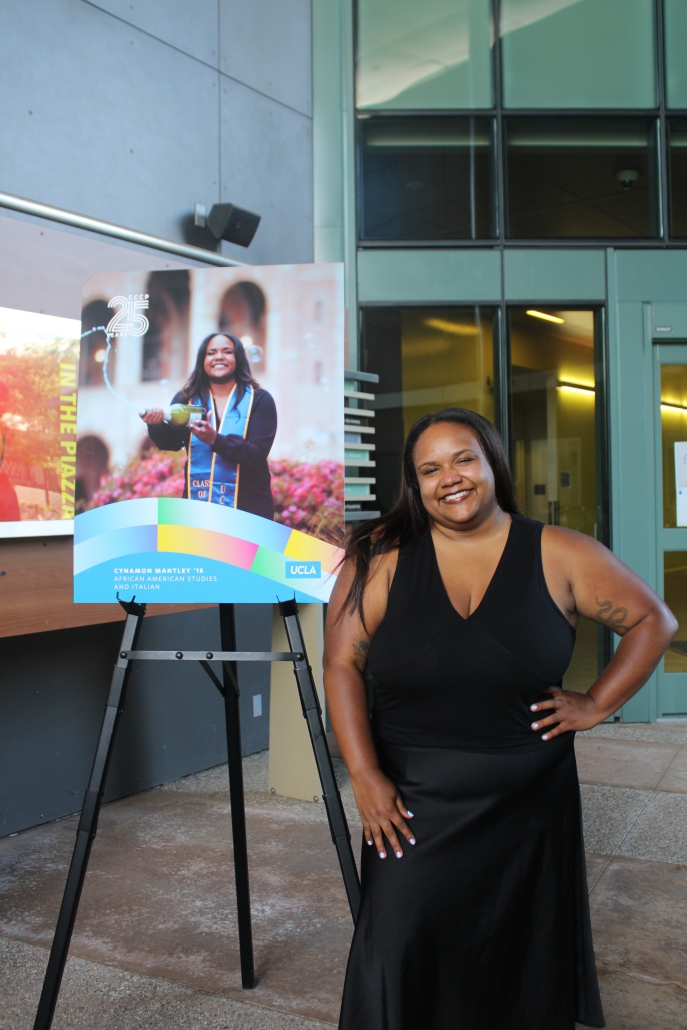 Cynamon Mantley, wearing a black sleeveless dress, poses with her hands on her hips next to a poster on an easel showing her UCLA graduation photo.