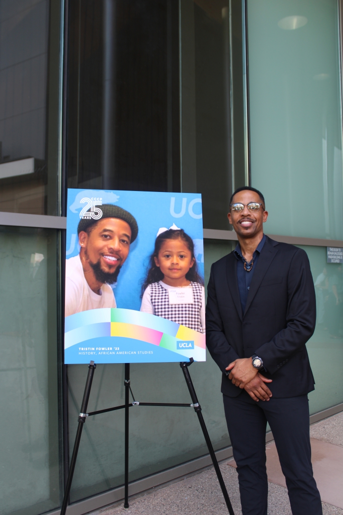 Tristin Fowler, wearing a dark suit, stands smiling next to a poster on an easel that displays a photo of him with his young daughter.