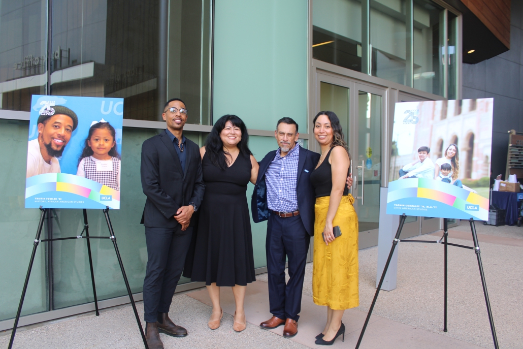: Four people stand together outside glass doors framed by two easel-mounted posters featuring UCLA alumni portraits.