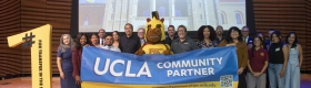 A group of people stands on stage holding a blue banner that reads "UCLA COMMUNITY PARTNER". A mascot stands in the center, and a large yellow cutout on the left reads "#1 FOR TRANSFER IN THE STATE / PASADENA CITY COLLEGE". A presentation screen behind them shows Royce Hall at UCLA along with UCLA and PCC logos.