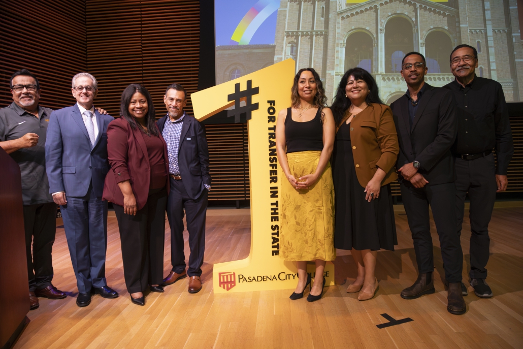 Seven people stand on stage posing on both sides of a large yellow cutout shaped like a number one, which reads "#1 FOR TRANSFER IN THE STATE" above the Pasadena City College logo.