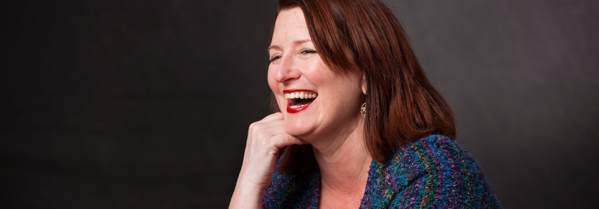 Katherine Miller, wearing gold bracelets and a colorful knitted shawl, rests her chin on one hand while laughing against a dark gray background.