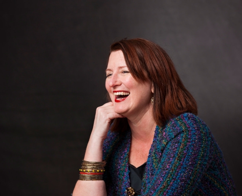 Katherine Miller, wearing gold bracelets and a colorful knitted shawl, rests her chin on one hand while laughing against a dark gray background.